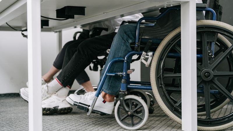 unter einem Schultisch sieht man, dass Schüler*innen sowohl auf Stühlen als auch im Rollstuhl sitzen